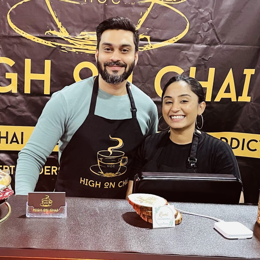 Two people wearing aprons in front of a 'High on Chai' branded backdrop.